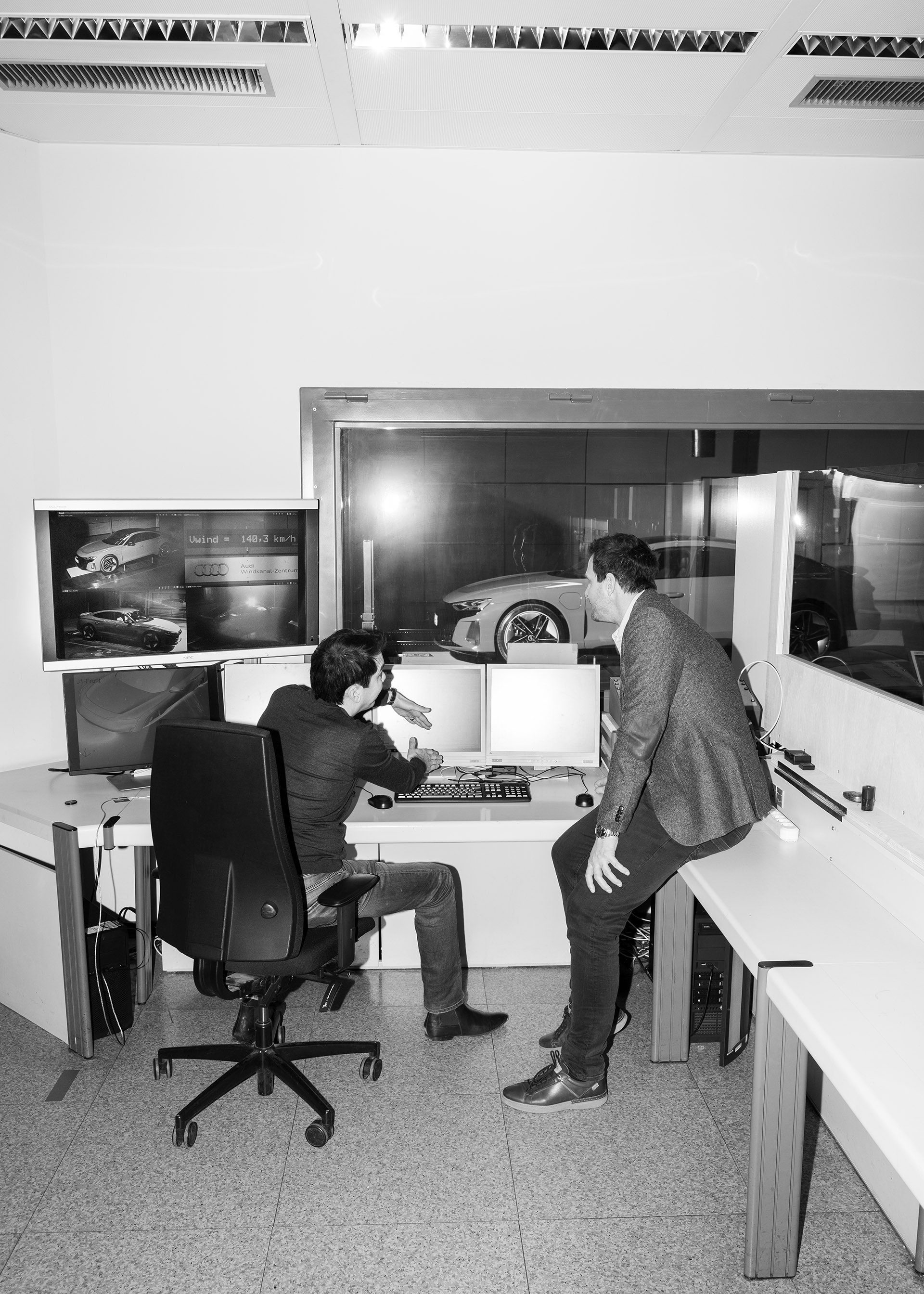 Kentaro Zens og Thomas Redenbach diskuterer foran flere dataskjermer med Audi RS e-tron GT stående bak et vindu i bakgrunnen.